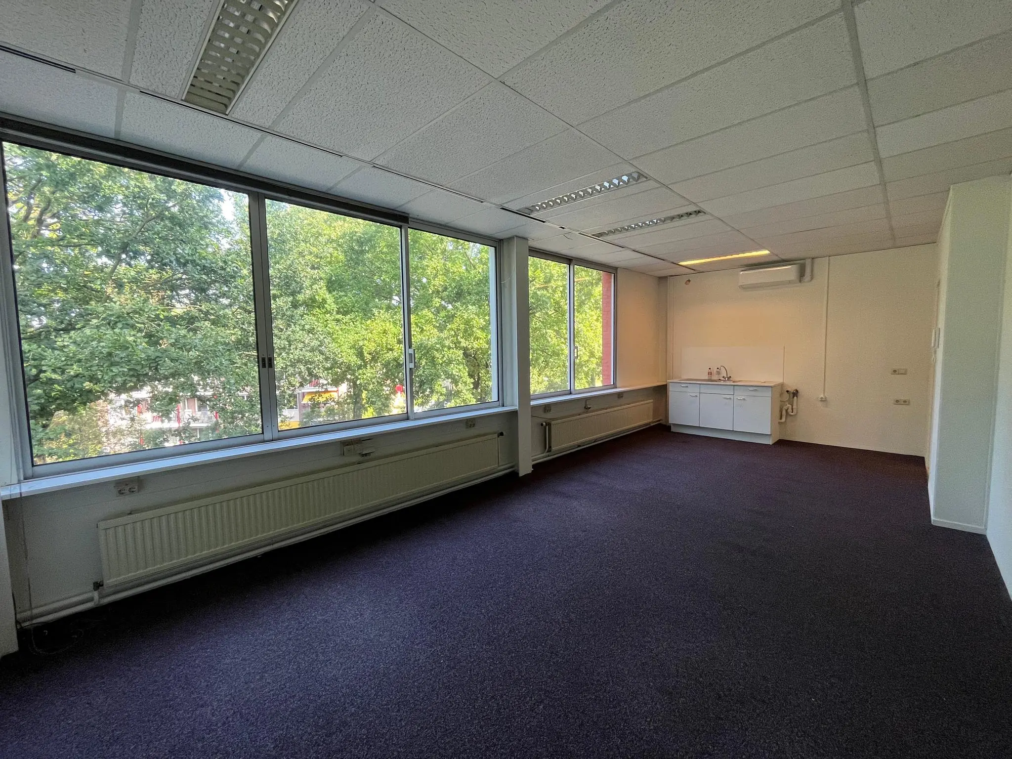 Lichte kantoorruimte aan de Jan van Eycklaan met grote ramen, paarse vloerbedekking en een kitchenette.